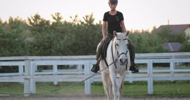 Sakin bir manege içinde görkemli beyaz bir ata binen kadın binici, sürücünün ve hayvanın arasındaki derin bağlantıyı vurgulayan sakin bir günbatımı antrenmanının keyfini çıkarıyor.