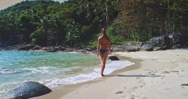 Bir kadın kumlu bir sahilde yürüyor, hafif dalgalar ayaklarına vuruyor. Bereketli yeşillik ve uzak tepeler sakin bir atmosfer yaratır, rahatlamak için mükemmel..