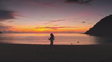 Tayland 'da pitoresk bir plajda koşuşturan siluetli sporcu kadın, çarpıcı bir günbatımının canlı renklerini kucaklarken özgürlüğü ve sağlıklı bir yaşam tarzını kutluyor.