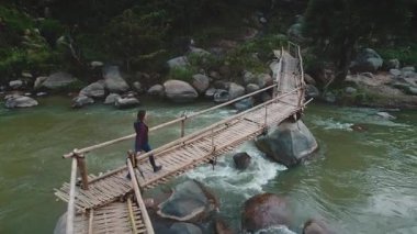Kişi, yemyeşil ve büyük kayalarla çevrili akan bir nehri kapsayan taşralı ahşap bir köprüden geçer. Sahne, doğal ortamdaki huzurlu bir anı yakalar.