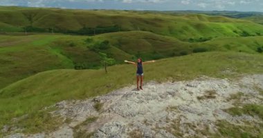 Genç bir kadın tepenin üstünde kolları açık duruyor, yuvarlanan yeşil tepelerin güzelliğini kucaklıyor ve bulutlu bir gökyüzünün altında Sumba Adası 'nın yemyeşil bitki örtüsünü kucaklıyor.