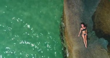 Bir insan, turkuaz suyla çevrili büyük bir kayanın üzerinde güneşlenir, açık bir günde güneşi emer. Dalgalar yavaşça kıyıya çarpar ve sakin bir atmosfer yaratır..