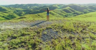 Endonezya 'nın Sumba Adası' nda özgürlüğünü kucaklayan genç bir kadın turist, kollarını açmış yemyeşil tepelerden oluşan cennet gibi bir manzarada gün batımının sıcacık parıltısıyla güneşleniyor.