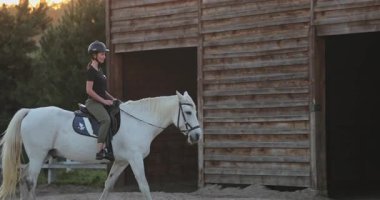 Dişi binici kask takıyor ve güzel beyaz bir ata biniyor. Ahırdan çiftlikte ayrılıp açık hava ve at arkadaşlığı özgürlüğünün tadını çıkarıyor.