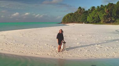 Genç bir kadın, köpeğiyle el değmemiş, tropik bitki örtüsü ve turkuaz sularla dolu, el değmemiş bir kumsalda, rahatlatıcı bir yürüyüşün tadını çıkarıyor.