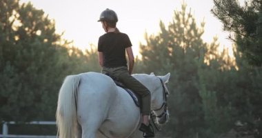 Genç bir kadın ormanda beyaz bir ata biniyor, ata binerken gün batımının tadını çıkarıyor, binicilik turizmini ve doğayla bağlantısını destekliyor.