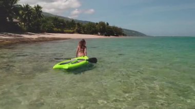 Kadınlar kumsalda güneşli bir günün tadını çıkarırlar. Turkuaz suda palmiye ağaçları ve okyanus dalgalarıyla çevrili bir sörf tahtasıyla yürürler.