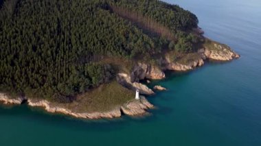 Kuzey İspanya 'nın resimli manzarası. Deniz feneri kayalıklarda izole edilmiş, orman ve tepelerle çevrili. Önlerinde turkuaz renkli deniz var. İHA ileri alçalıyor ve kamerayı aşağı eğiyor