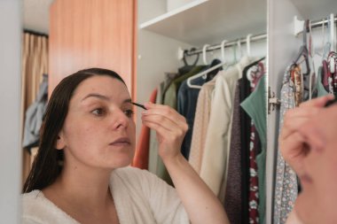 Beautiful Hispanic woman in a white bathrobe applying black makeup to her eyebrows with a small brush in front of the mirror on the closet door in her bedroom