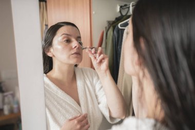 Beautiful Hispanic woman in a white bathrobe combing her eyebrows in front of the mirror on her closet door in her bedroom