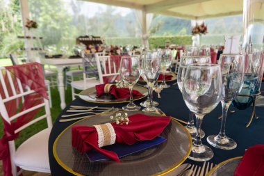 Bahçede bir parti masası, kırmızı peçetelerle süslenmiş, mavi masa örtüsü ve arka planda beyaz sandalyeler bulunan kristal bardaklar.