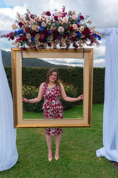 Beautiful young Latin woman wearing a red dress, standing in a garden posing behind an antique gold frame decorated with a bouquet of roses at a party during the day
