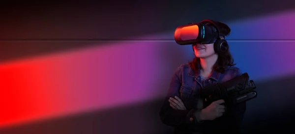 年轻美丽的惊慌失措的女人站在那里 手里拿着虚拟现实眼镜和游戏枪 背靠着墙 蓝光和紫光 — 图库照片