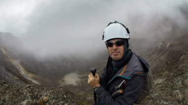 Genç dağcı, bulutlu bir günde, Guagua Pichincha volkanının yanında tek başına yürüyüş direğini kullanarak siyah ceketli ve kasklı yürüyor.