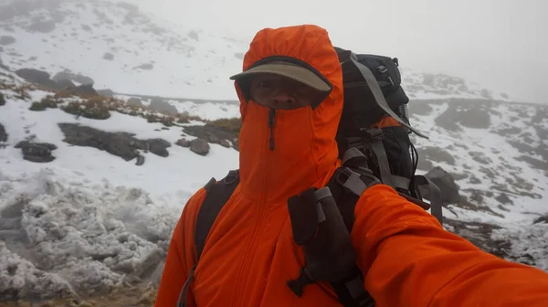 Young climber man with backpack, red coat and cap taking a selfie while climbing a snowy mountain on a cloudy day
