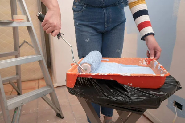 Close up on standing woman dressed in overalls and striped blouse, seen from front, dipping a roller in the tray with blue paint, inside an empty room with white walls half painted with blue paint