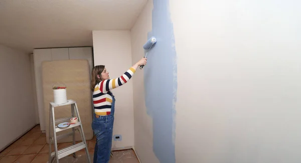 Uma linda designer de mulher faz reparos pinta uma parede cinza com tinta  verde com um pincel camisa xadrez cabelo comprido e jeans reparo no  apartamento