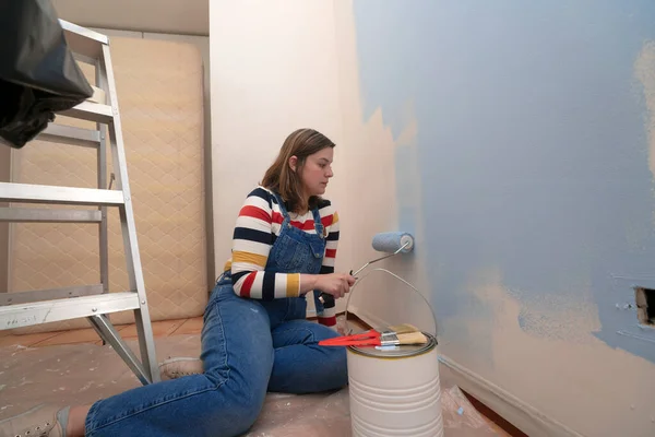 Kneeling woman dressed in overalls and striped blouse, profile view, painting a white wall with a roller with blue paint, inside an empty room with a metal ladder, paint jar and brushes