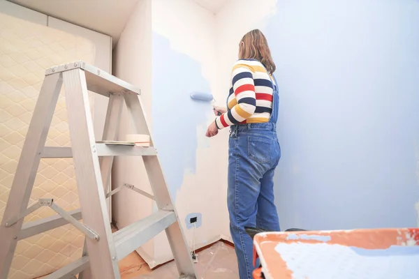Standing woman dressed in overalls and striped blouse, seen from behind, painting a white wall with a roller with blue paint, inside an empty room with a metal ladder