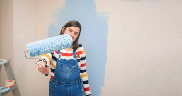 Close-up of roller with blue paint in the hand of a standing woman dressed in overalls and striped blouse, seen from the front, against background of half-painted white wall with strokes of blue paint