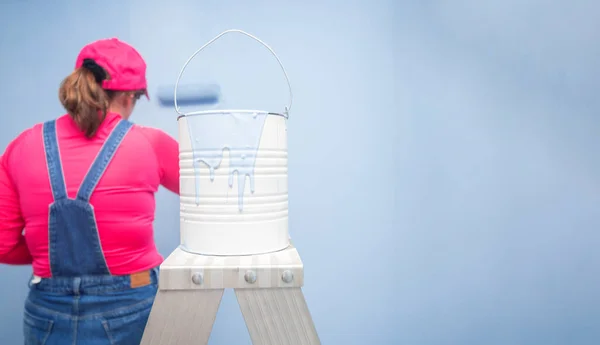 Close-up of a white paint can with blue paint dripping on a metal ladder, with a background of a woman dressed in overalls and a red t-shirt painting with a roller a blue wall in an empty room