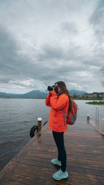 Turuncu ceketli, sırt çantalı güzel Latin Amerikalı bir kadın bulutlu bir günde dağlarla çevrili bir göldeki ahşap iskelede fotoğraf çekiyor.