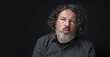 Beyaz sakallı, siyah kıvırcık saçlı, şüpheli bir şekilde solda görünen, siyah arka planda siyah gömlekli Latin bir adamın portresi.
