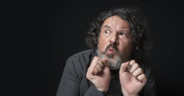 Beyaz sakallı, siyah kıvırcık saçlı, korkmuş, siyah arka planda siyah gömlek giyen Latin bir adamın portresi.