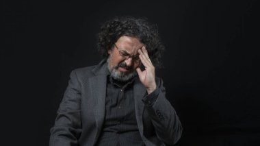 Beyaz sakallı, siyah kıvırcık saçlı, eli alnında, siyah arka planda siyah gömlekli Latin bir adamın portresi.