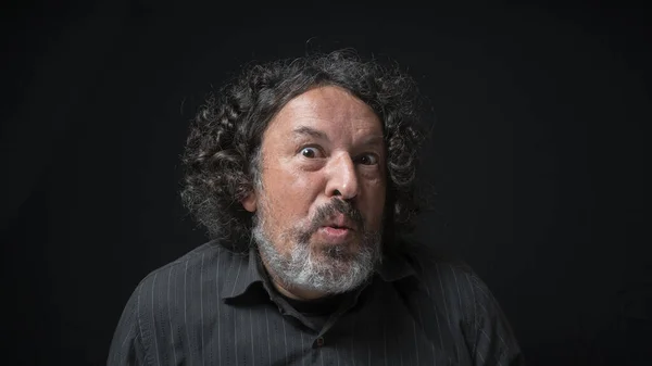 Portrait of latin man with white beard and black curly hair with funny expression, grimacing with eyes and mouth, wearing black shirt against black background