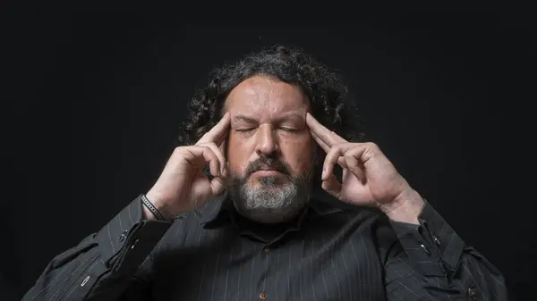 Portrait of latin man with white beard and black curly hair with concentrated expression, with his fingers on his temples in his hand, wearing black shirt against black background