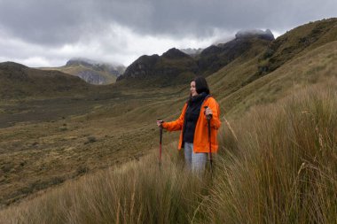 Kırmızı ceketli güzel bir kadın bulutlu bir günde arka planda dağlarla çalıların ortasında yürüyüş direkleri ile yürüyor.