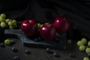 Kırmızı elmalar, yeşil üzümler ve siyah seramik bir masanın üzerinde siyah kumaşla doğranmış yaban mersinleri.