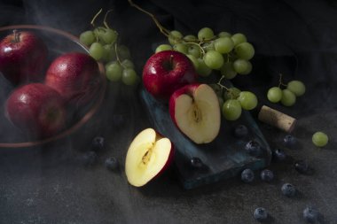 Kırmızı elmaların, yeşil üzümlerin ve etrafındaki buharlı yaban mersinlerinin siyah seramik bir masanın üzerindeki kesme tahtasının üst görüntüsü.