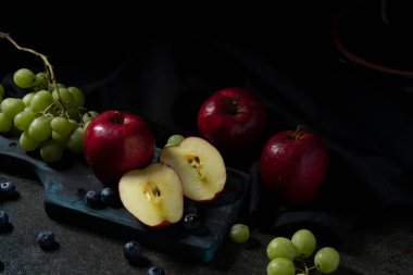 Arka planda siyah bez olan siyah seramik bir masanın üzerinde yarım kalmış bir elma, kırmızı elma, yeşil üzüm ve yaban mersini yakın plan.