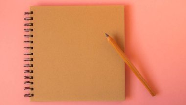 Açık pembe arka planda, kapağında kalem olan not defteri.