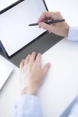 Mavi gömlekli orta yaşlı bir kadının elleri beyaz masada tablet ve dizüstü bilgisayarla çalışıyor.