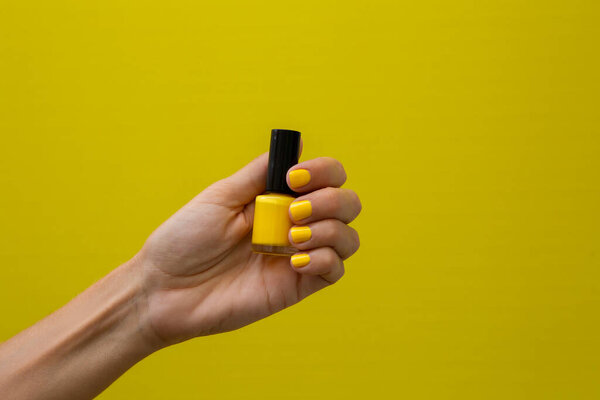 Горизонтальная фотография женской руки с желтыми ногтями, держащими бутылку желтого лака для ногтей на желтом фоне.