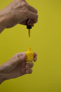 Vertical photograph of the drop of yellow nail polish falling on the bottle while being held by a woman's hand on a yellow background. clipart