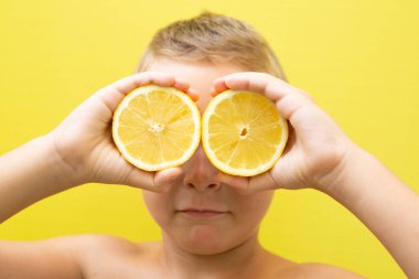 Sarı saçlı 7 yaşında bir çocuk. Sarı arka planda gözlerini iki limon kapamış.