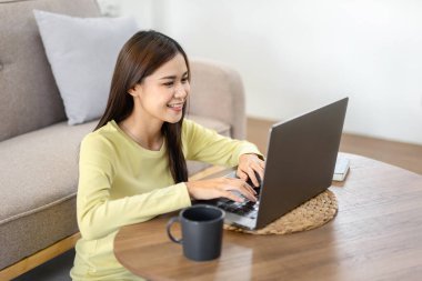 Genç bir kadın yerde oturmuş gülümsüyor ve iş raporlarını yazmak için dizüstü bilgisayar kullanıyor. Çalışırken ve internetten alışveriş yaparken evdeki oturma odasında dinleniyor..