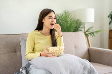 Genç Asyalı kadın yastığa bir kase patlamış mısır koyup patlamış mısır yerken büyük rahat koltukta oturup evdeki oturma odasında televizyon seyrediyor..