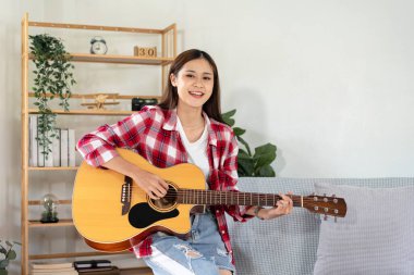 Genç kadın, evdeki oturma odasında dinlenirken şarkı söylemek ve pop şarkılarıyla gitar çalmak için temel gitar akorları çalışıyor..