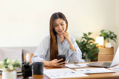 Serbest çalışan kadın akıllı telefondan iş verilerini okuyor ve merkez ofisin oturma odasındaki masada çalışmak için dizüstü bilgisayar kullanıyor..