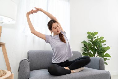 Genç kadın, evindeki oturma odasında yoga yaptıktan sonra koltukta rahatlamak için kollarını esnetiyor..