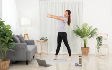 Genç bir kadın laptopta yoga dersi alıyor ve evinde kollarını mindere uzatarak yoga egzersizi yapıyor..