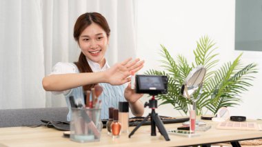 Genç güzellik etkileyicisi, sosyal medyada veya stüdyoda web blogcusu paylaşımı için kameraya video kaydederken ve kozmetik ürünlerini test etmek ve tavsiye etmek için ruj ürünü gösteriyor.