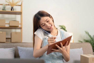 Genç girişimci akıllı telefondan müşteriyle konuşuyor ve bilgisayardaki siparişleri kontrol ediyor rahat koltukta otururken ve ofisteki oturma odasında çalışırken dizüstü bilgisayara veri yazmak için..