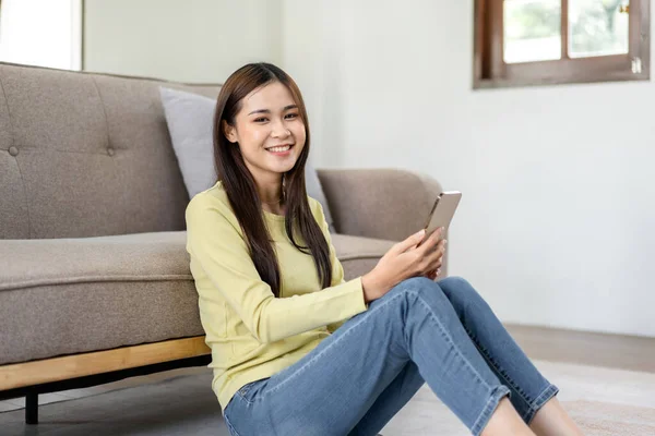 年轻女性正坐在地板上 靠着舒适的沙发 一边在家里客厅里工作 一边用智能手机查看商业数据 — 图库照片