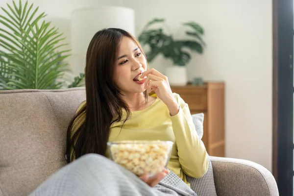 若いですアジアの女性を持っていますボウルのポップコーンを食べますポップコーンながら横になっていますザ大快適なソファへ見ます映画上のテレビで自宅のリビングルーム — ストック写真